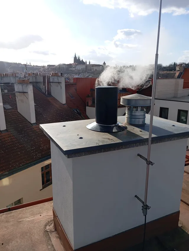 Kouřící komínové vyústění na střeše pražského domu, v pozadí panorama Hradčan